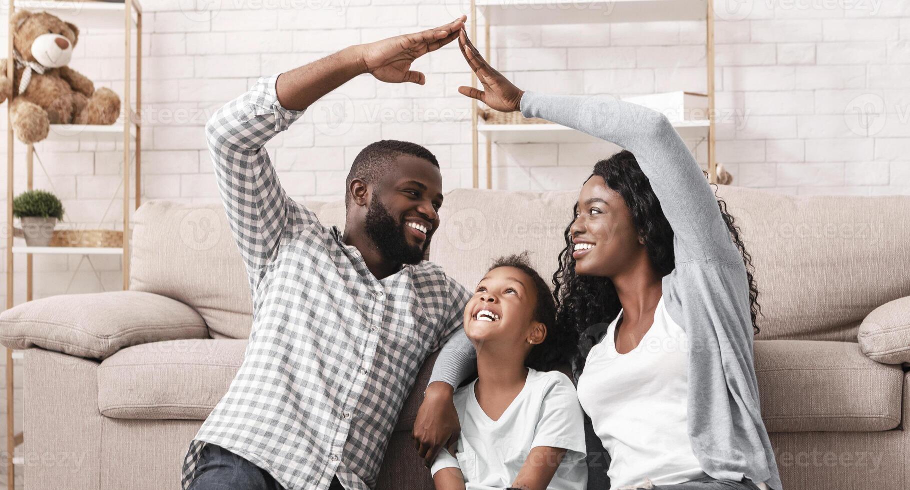 Family Care And Protection Concept. Black parents making symbolic roof of hands above their little daughter, sitting on floor at home photo
