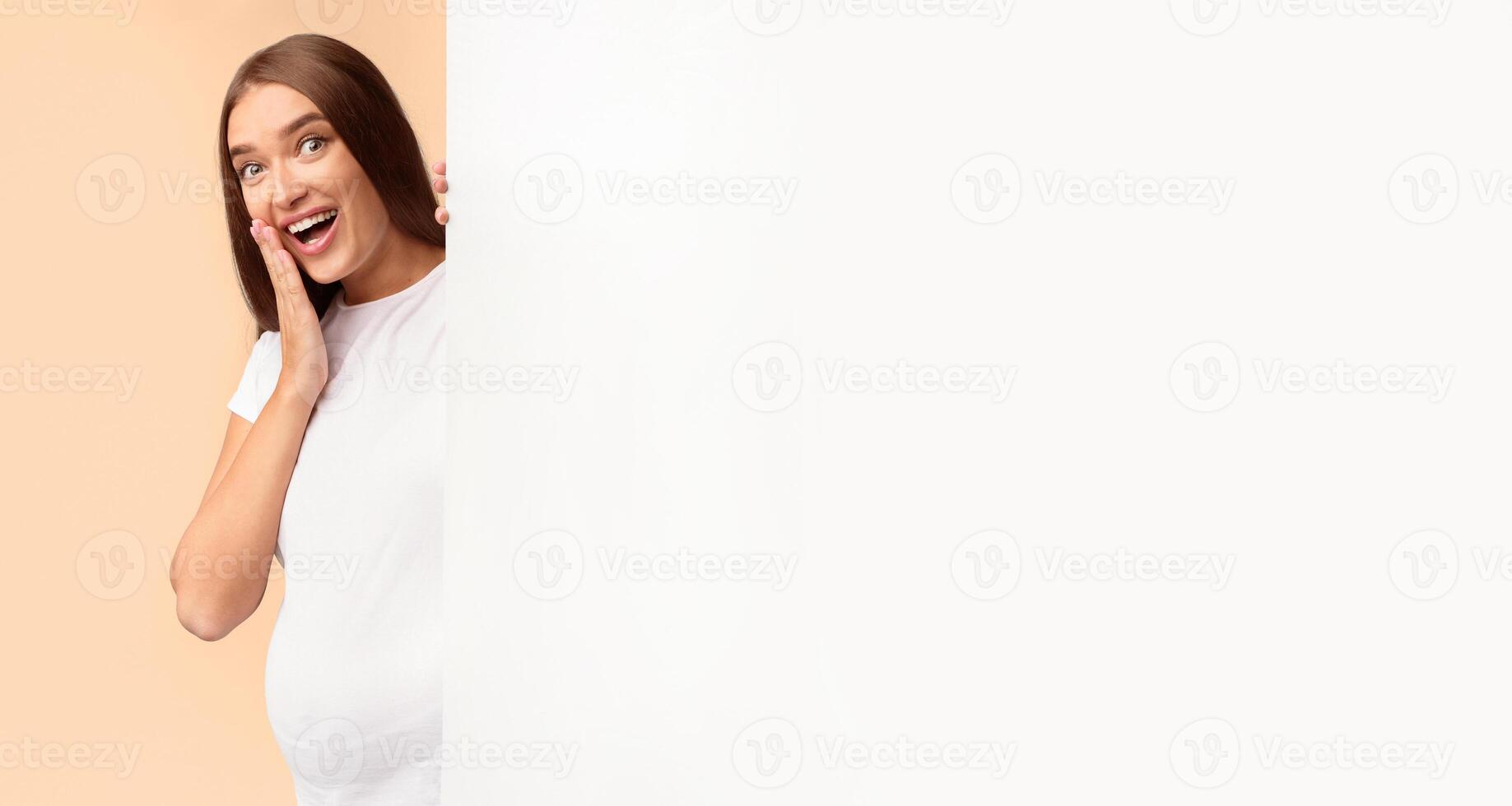 Great Offer. Excited Pregnant Girl Peeking Out Of Blank White Poster Standing Over Gray Studio Background, Mockup, Copy Space photo
