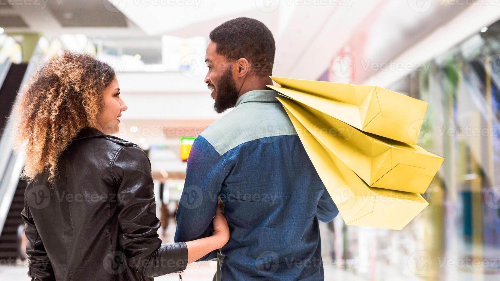 African American man and woman are standing in a busy mall, holding multiple shopping bags, enjoying their shopping trip together as they browse through various stores, back view photo