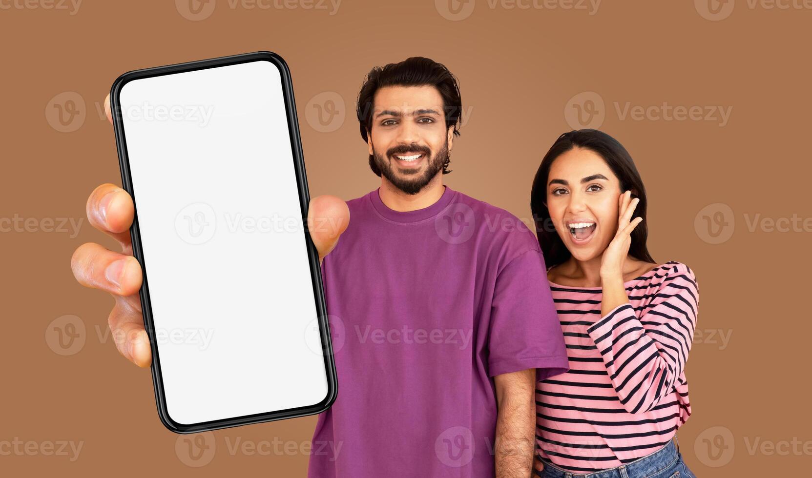 Multiracial man and woman are standing in front of a brown background. The man is wearing a purple t-shirt and holding a smartphone with a blank screen in front of him. photo