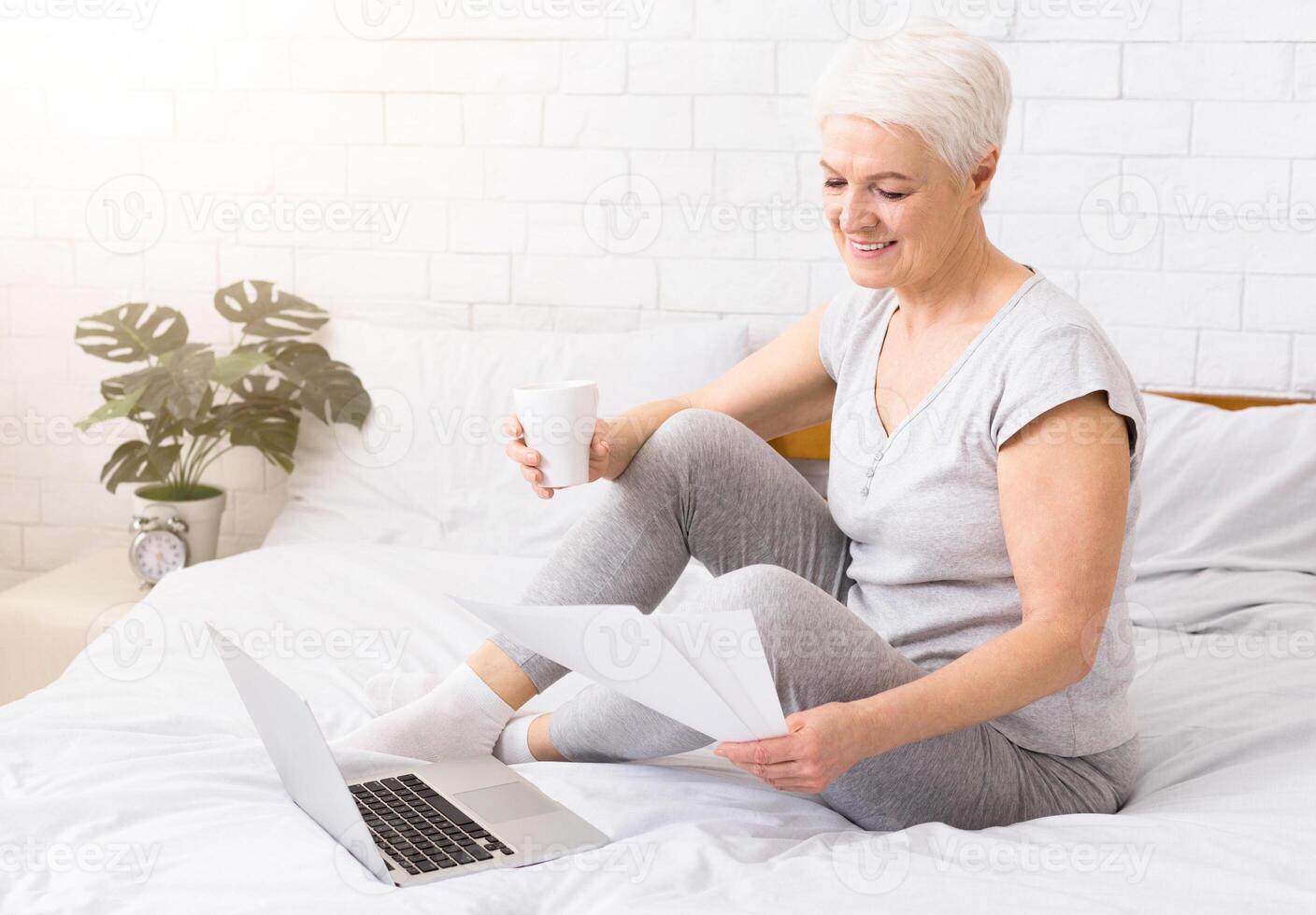 Senior business woman working with documents and laptop, sitting with coffee in bed, working from home, free space photo