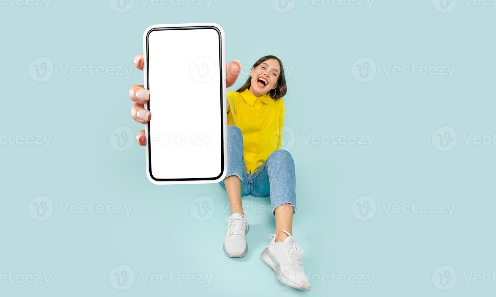 A woman is seated on the floor, holding a cell phone in her hands. She appears focused on the devices screen, possibly texting or browsing. The background is plain and simple. photo