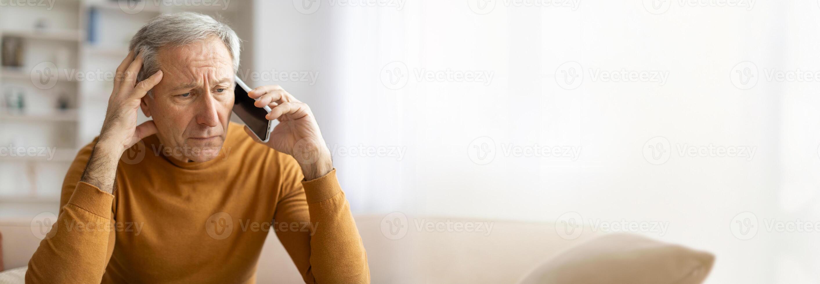 A senior man with gray hair sits on a couch in his living room, talking on the phone with a worried expression on his face. He appears concerned and is holding his hand to his forehead, copy space photo
