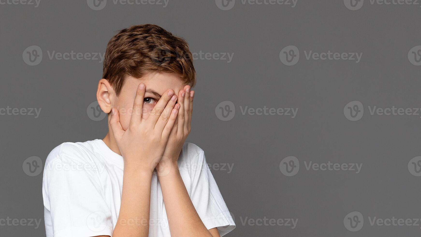 A young boy in a white t-shirt with a neutral background covers his face with his hands, expressing shame, fear, or shyness photo