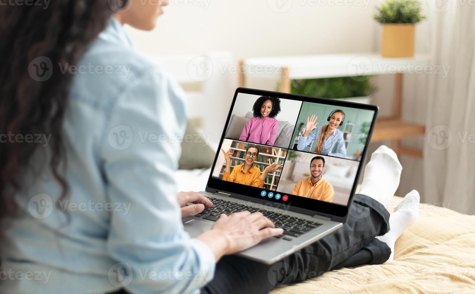 A woman sits on a bed in her home, using a laptop to chat with friends. She is smiling and typing on the keyboard. The laptop screen shows four people in different windows photo