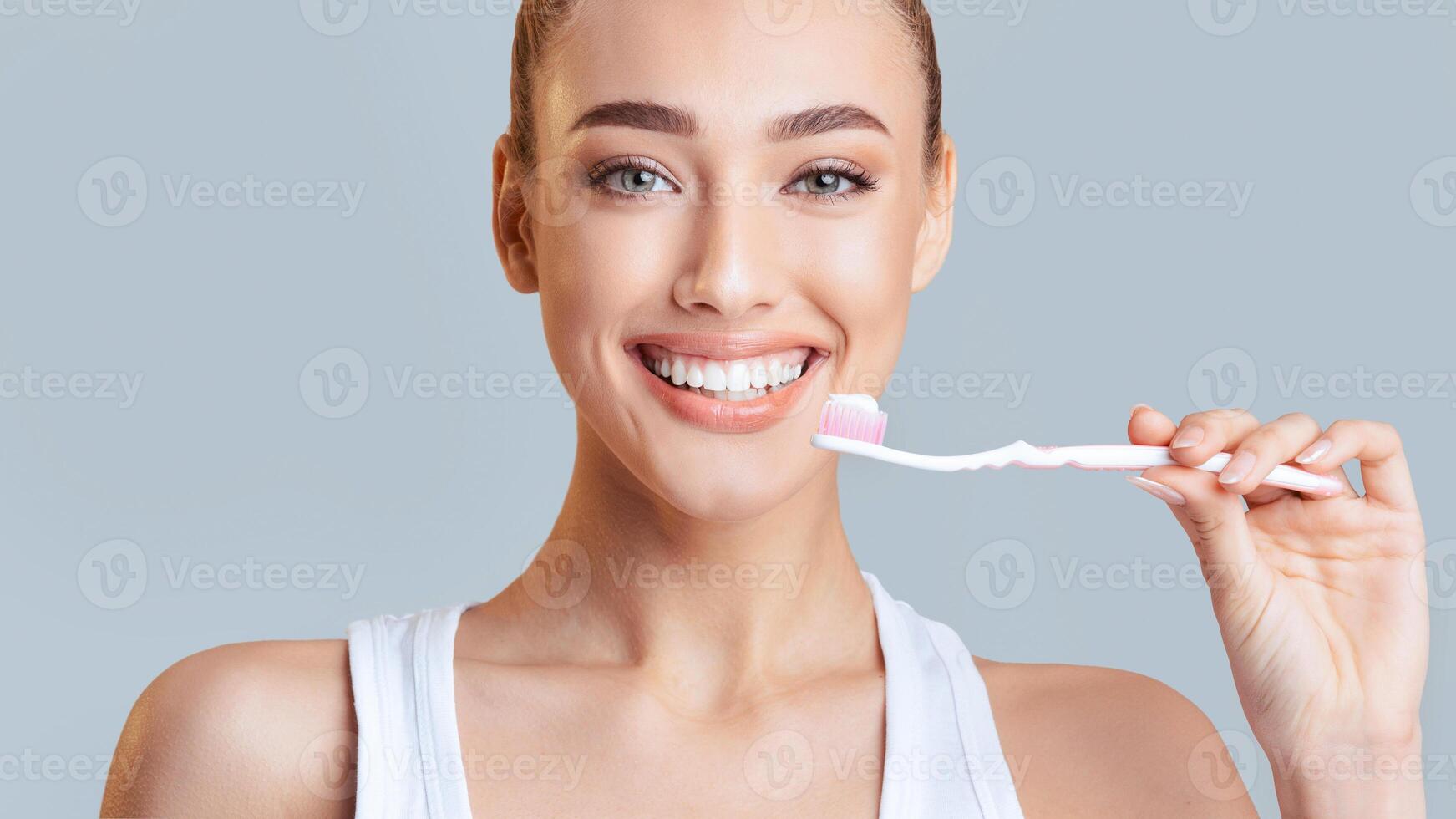 This image shows a woman with a white tank top brushing her teeth with a toothbrush and toothpaste. She has a bright smile and appears happy and healthy. photo