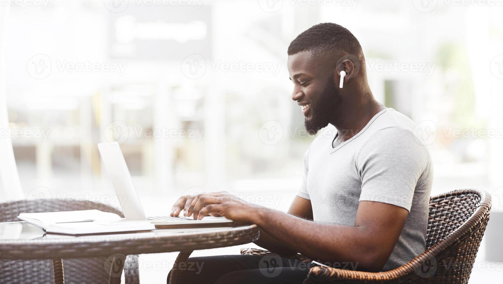 A smiling man works on his laptop while sitting at a table in a cafe. He is wearing earbuds and has a focused expression. photo