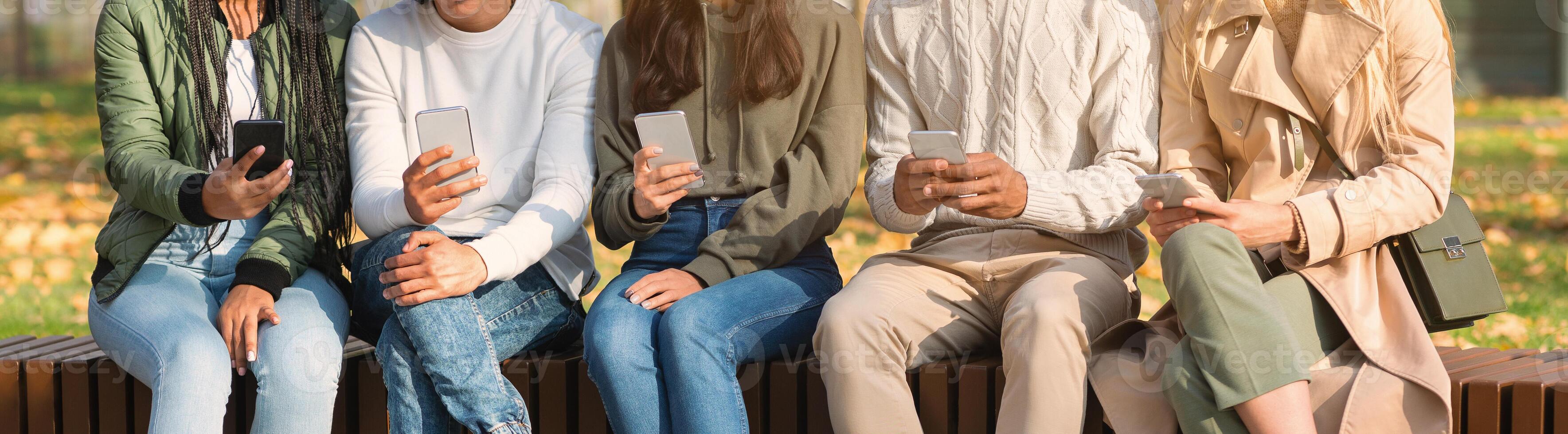 Close up of international young friends sitting in row on bench and using smartphones, panorama photo