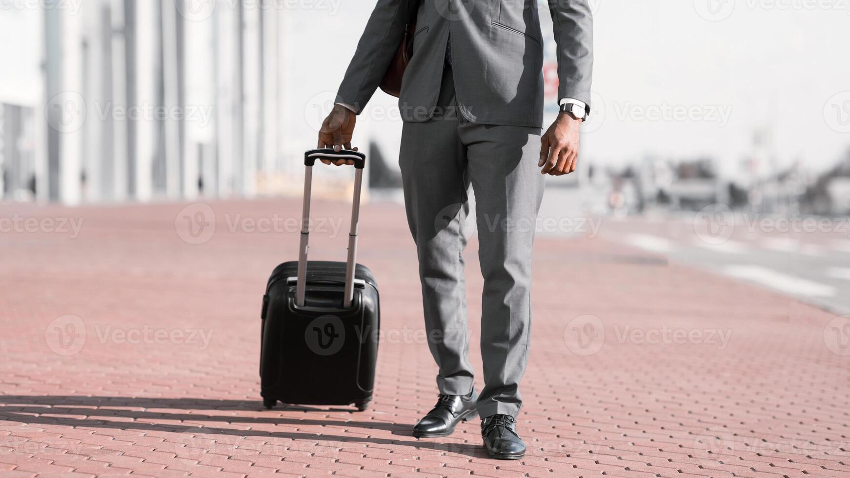 Unrecognizable black businessman walking with rolling suitcase outside a modern building, ready for a business trip or travel. photo
