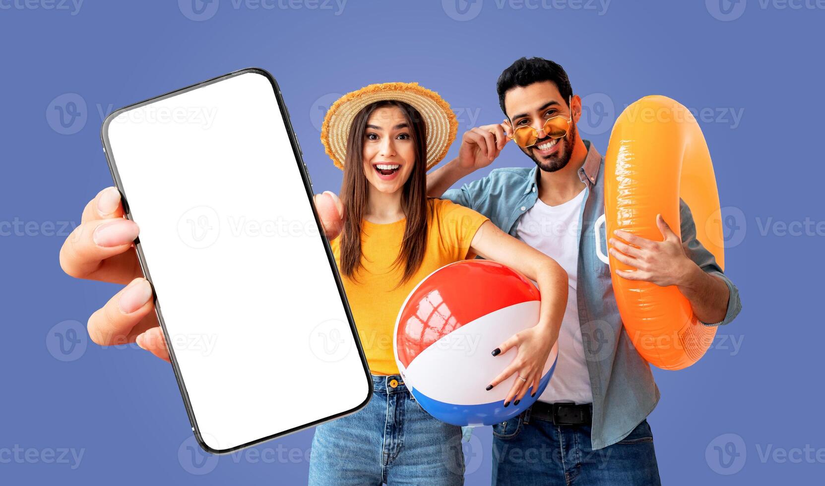 A young arabic couple is excited about their summer vacation. The woman is holding a beach ball and smiling, while the man is holding an inflatable ring. They are both pointing to a smartphone photo