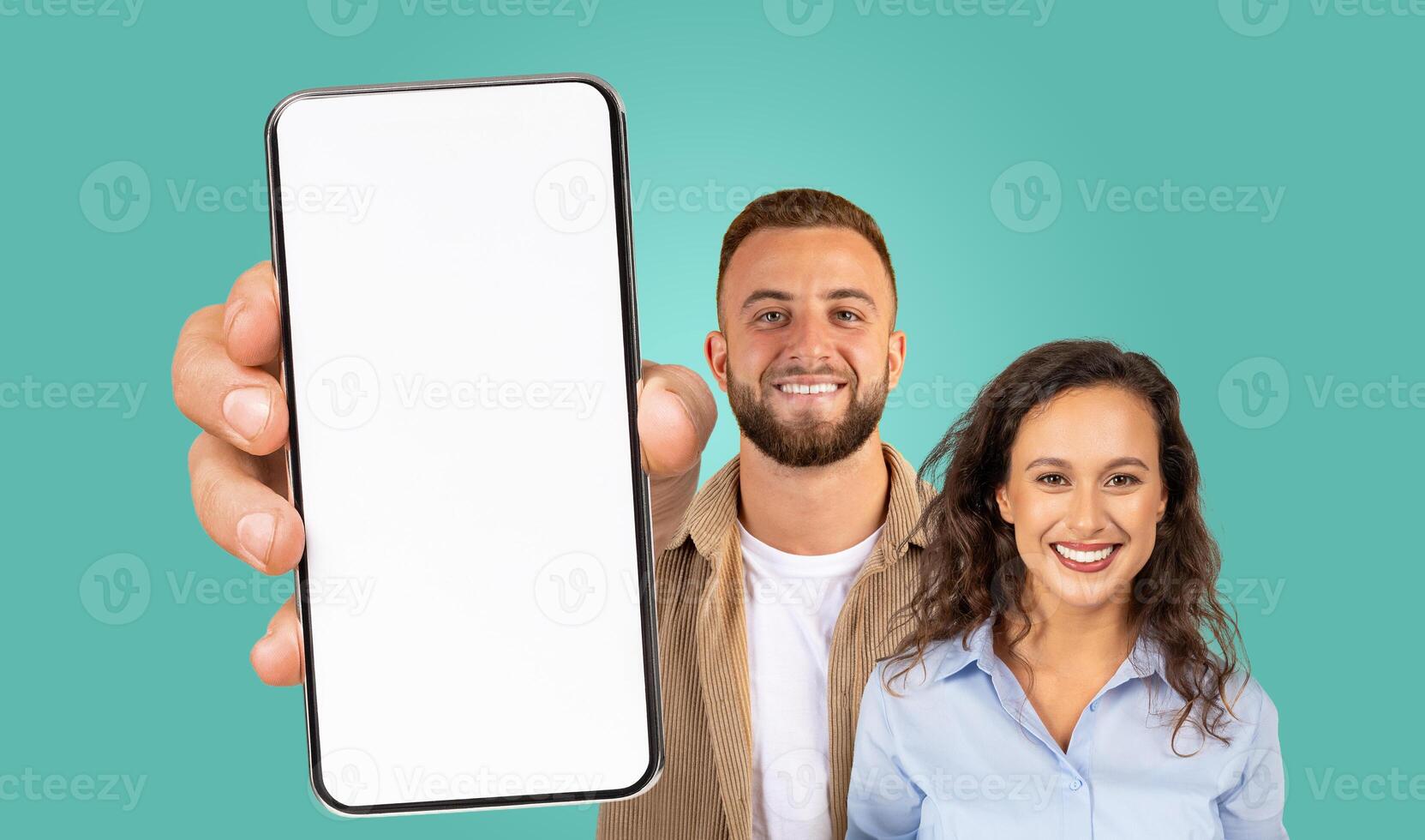 A man and a woman are standing together against a teal background. The man is holding a smartphone with a blank screen towards the camera. They are both smiling and looking directly at the camera. photo