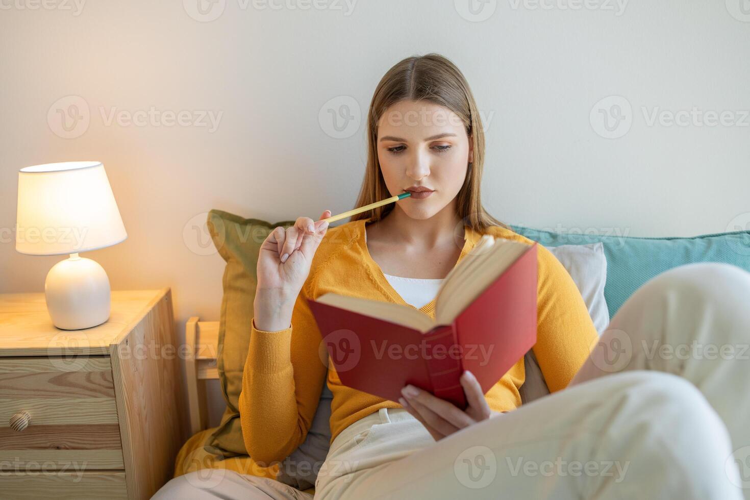 A girl is seated on a bed, engrossed in a book she holds in her hands. The room is well-lit, and she appears focused on the text as she reads. photo