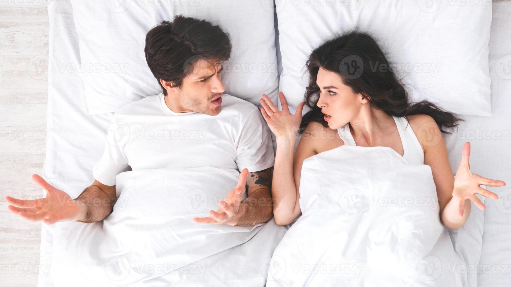 A young couple is lying in bed, arguing with each other. The man is lying on his back with his arms outstretched, while the woman is lying on her side, facing him. They both appear to be angry photo