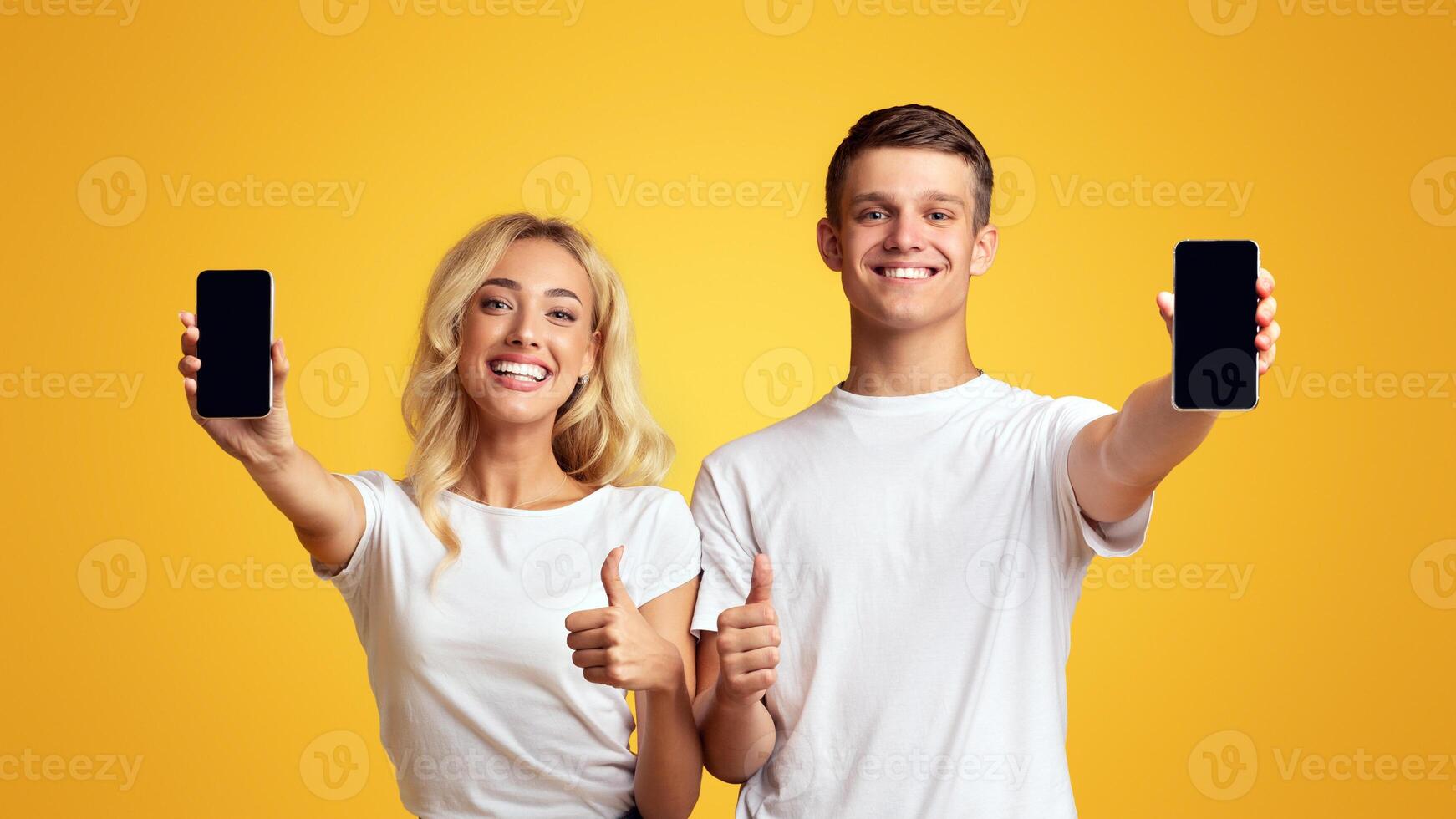 Cool application. Smiling young guy and girl demonstrating smartphones with blank screens and showing thumbs up, panorama with free space photo