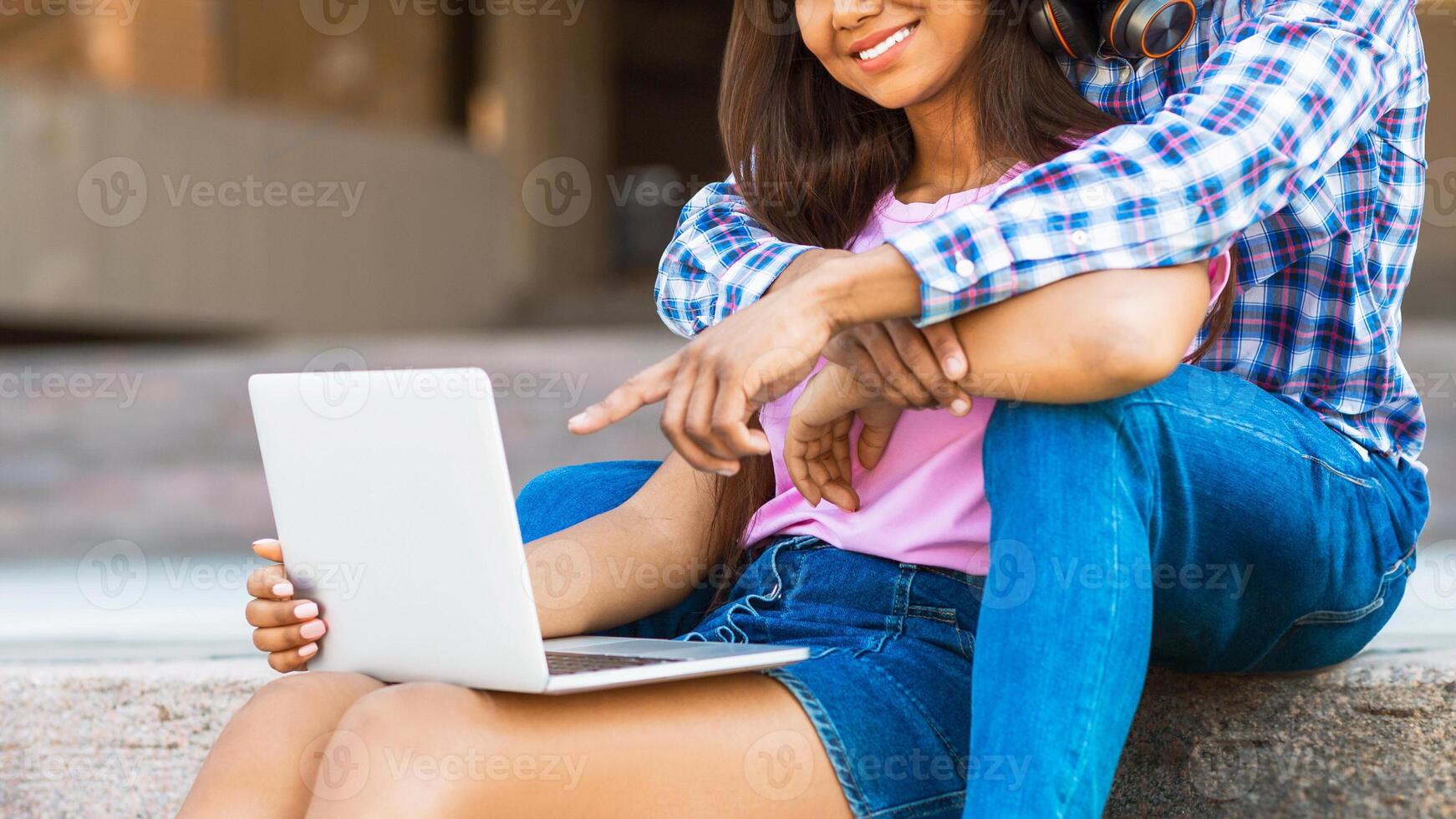 A teen black couple is sitting on concrete steps outside. The girl is using a laptop while guy wraps his arm around her shoulders and points at the screen, cropped photo