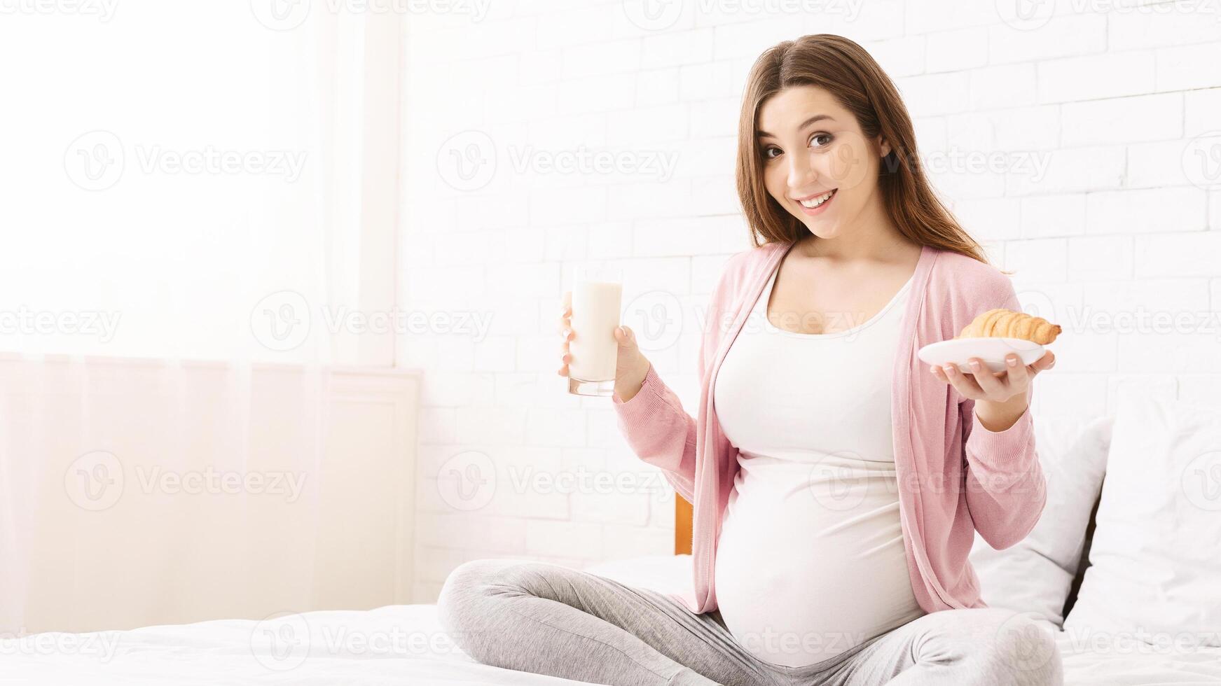 A pregnant woman is sitting comfortably in bed, smiling while holding a glass of milk and a pastry. She appears content and relaxed, showcasing a moment of calm and nourishment during her pregnancy. photo