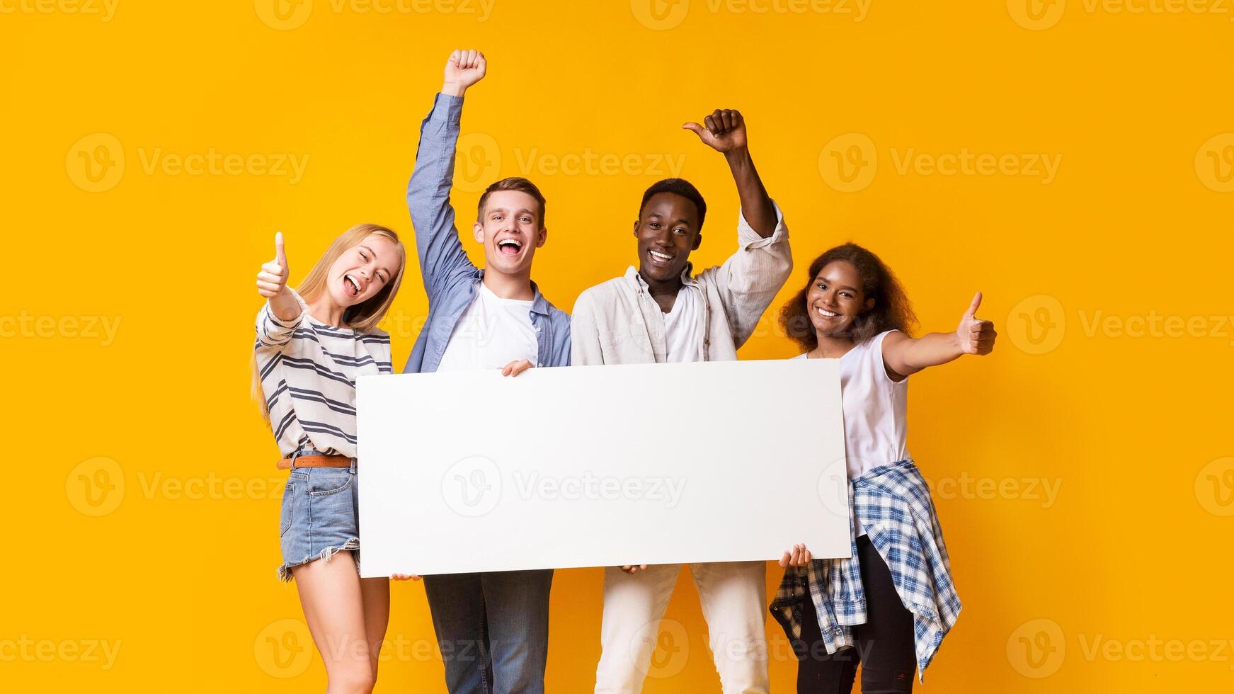 Cheerful international students with white empty board over yellow background photo