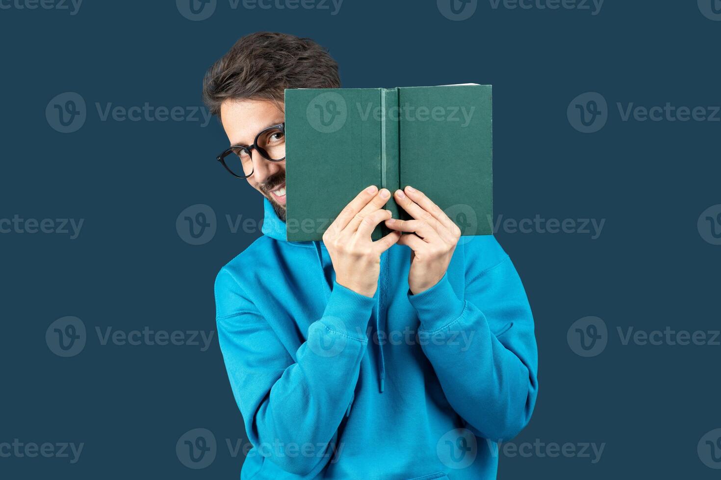 A young man with glasses smiles mischievously while holding a green book in front of face. He wears a blue hoodie and stands against a solid backdrop, creating a lighthearted and vibrant atmosphere. photo