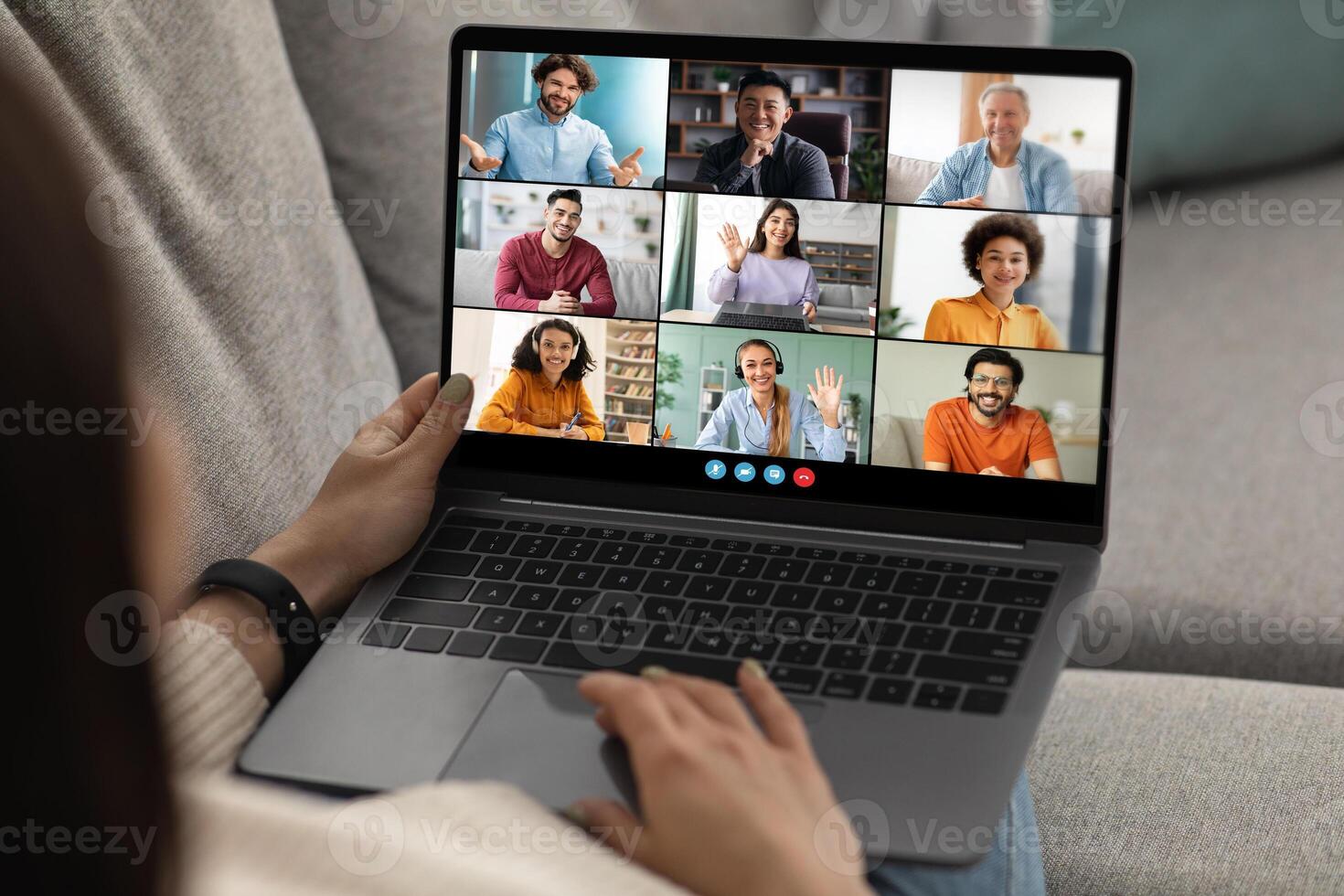 A woman sits on a couch and uses a laptop to participate in a virtual meeting. The laptop screen displays nine individual feeds of the other participants. photo