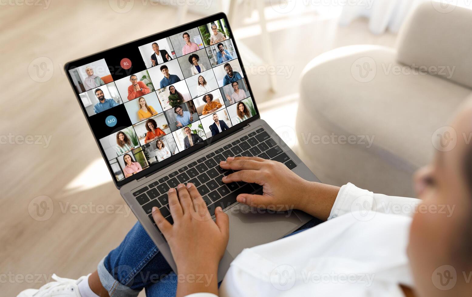 African American lady sits on a couch, holding a laptop in her lap. The laptop screen displays a conference with a grid of numerous participants. photo