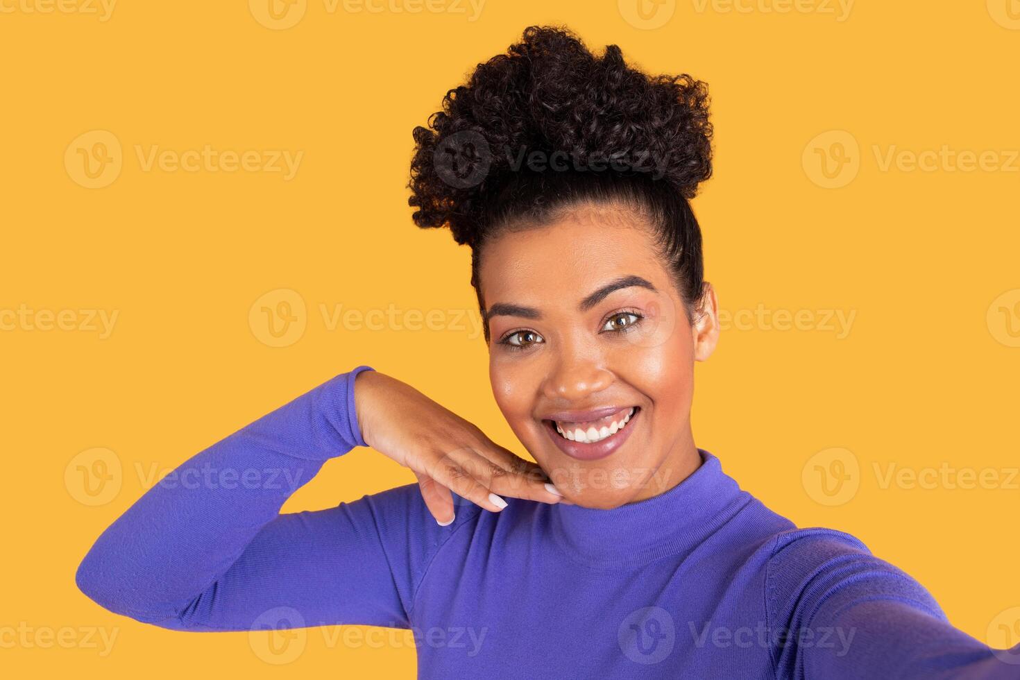 A woman with curly hair showcases a joyful expression while posing playfully against a vibrant yellow background. She wears a long-sleeved top and gestures with her hand under her chin photo