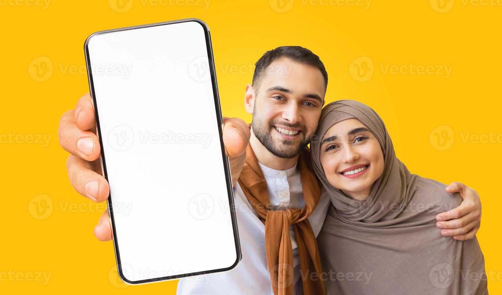 A young Muslim couple smiles at the camera while holding a smartphone with a blank screen. They are standing in front of a bright yellow background. The woman is wearing a hijab photo