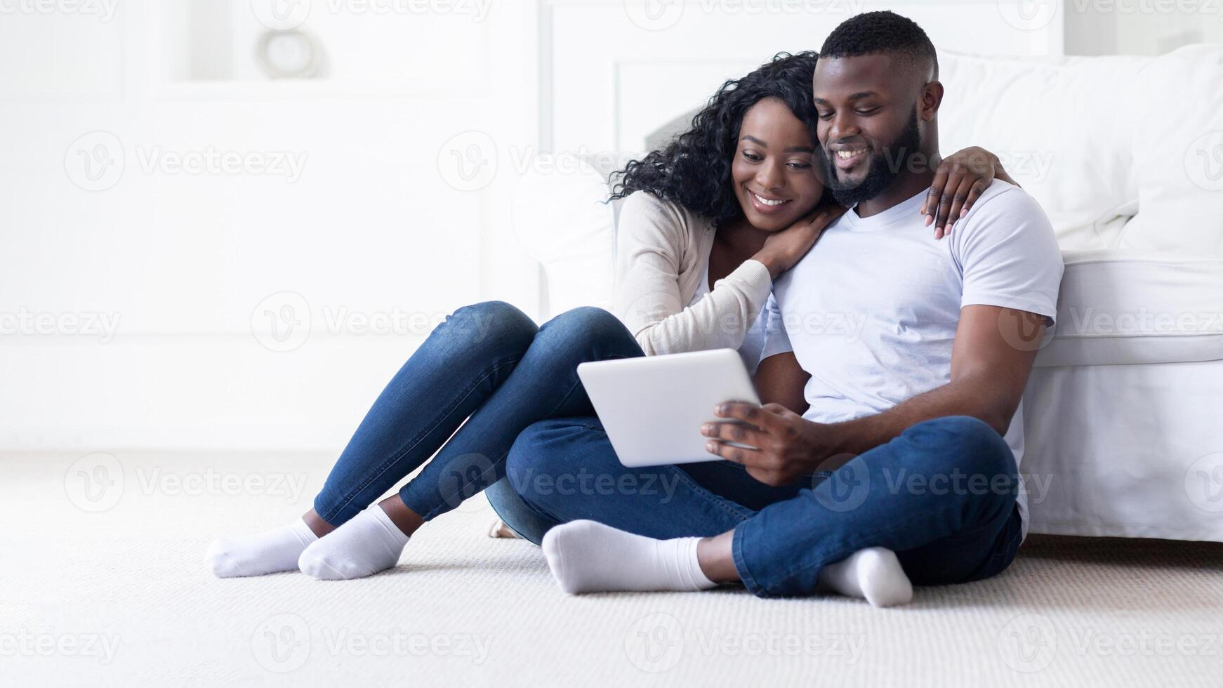 A couple sits on a carpeted floor, using a tablet device. The woman is resting her head on the mans shoulder, and they appear happy and relaxed. photo