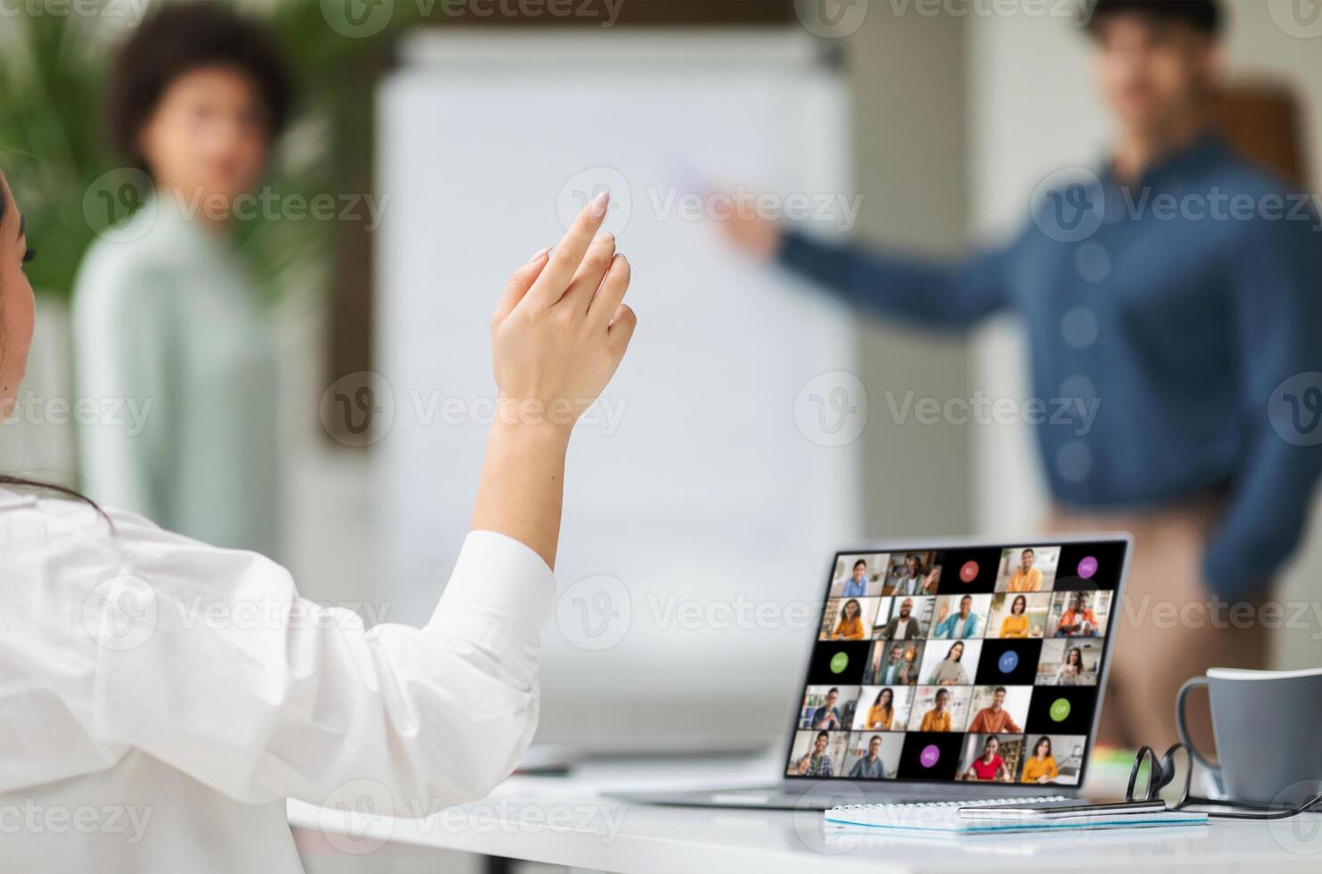Woman sitting in a meeting, raising her hand to ask a question. The room appears to be a hybrid setting, with the woman in person and other participants appearing on the laptop screen photo