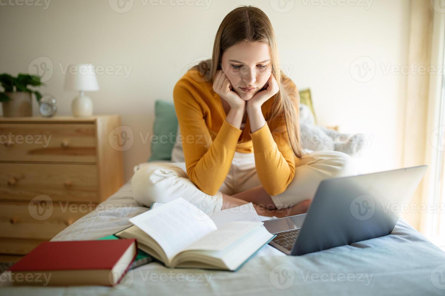 A teen girl sits on her bed in a cozy, well-lit bedroom. She is surrounded by open books and using a laptop, deeply focused on her work or studies. photo