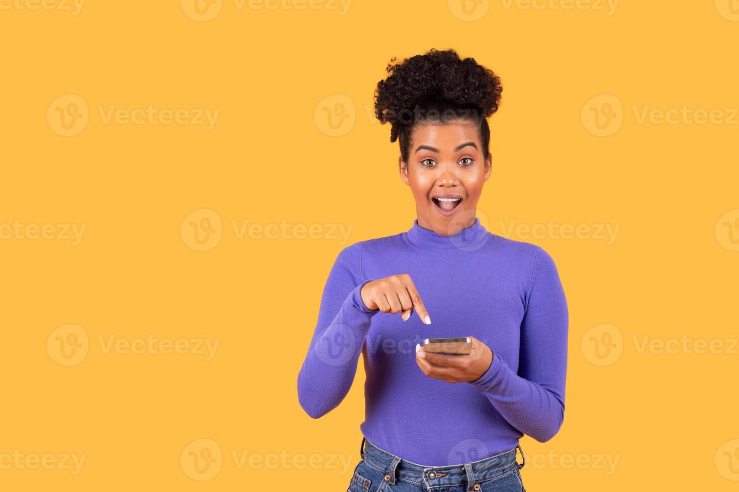 A young woman with curly hair wearing a purple top seems surprised while interacting with her smartphone. The vibrant yellow backdrop enhances her joyful expression, creating an energetic atmosphere. photo