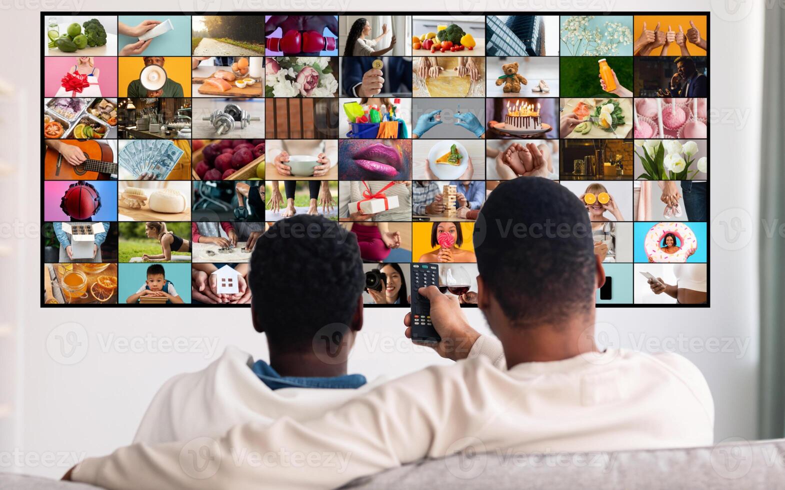 Two men are sitting on a couch and watching television. The television screen is showing a mosaic of various images, depicting different scenes and activities. photo