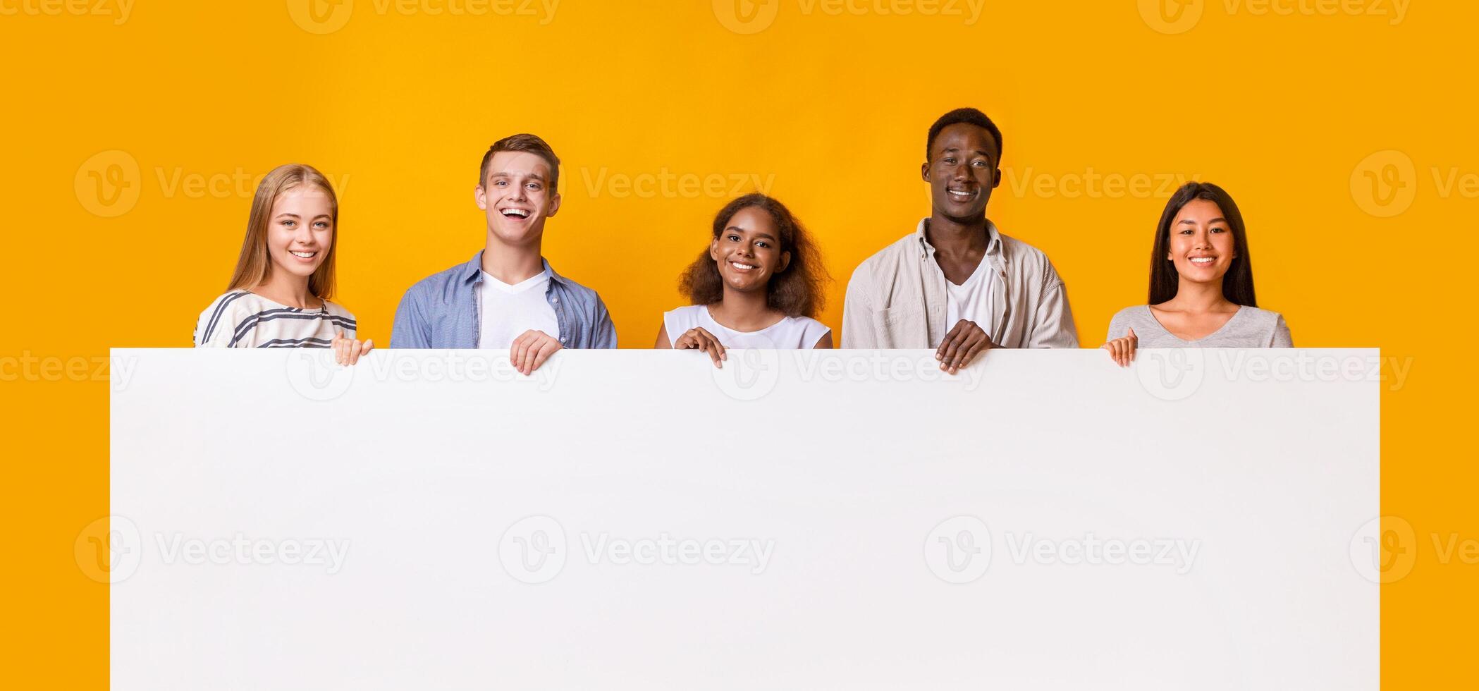 Smiling group of multiracial students standing together in row and displaying white empty placard, yellow studio background photo