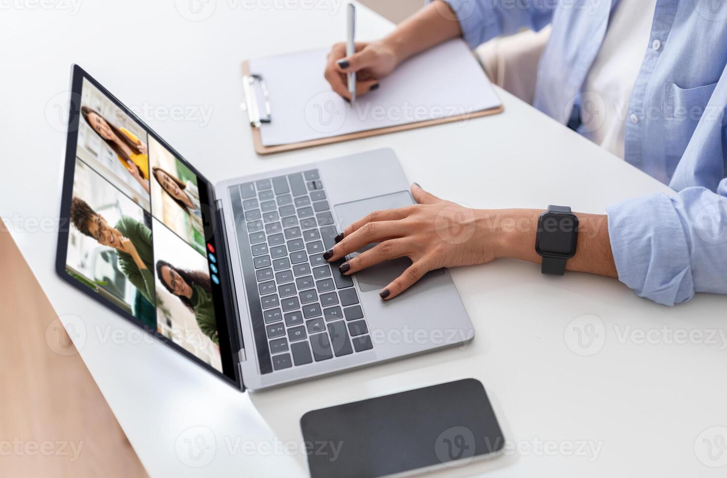 A woman is seated at a white desk with a laptop in front of her. She is using one hand to navigate the laptop while the other is resting on the desk. The laptop screen displays a conference photo