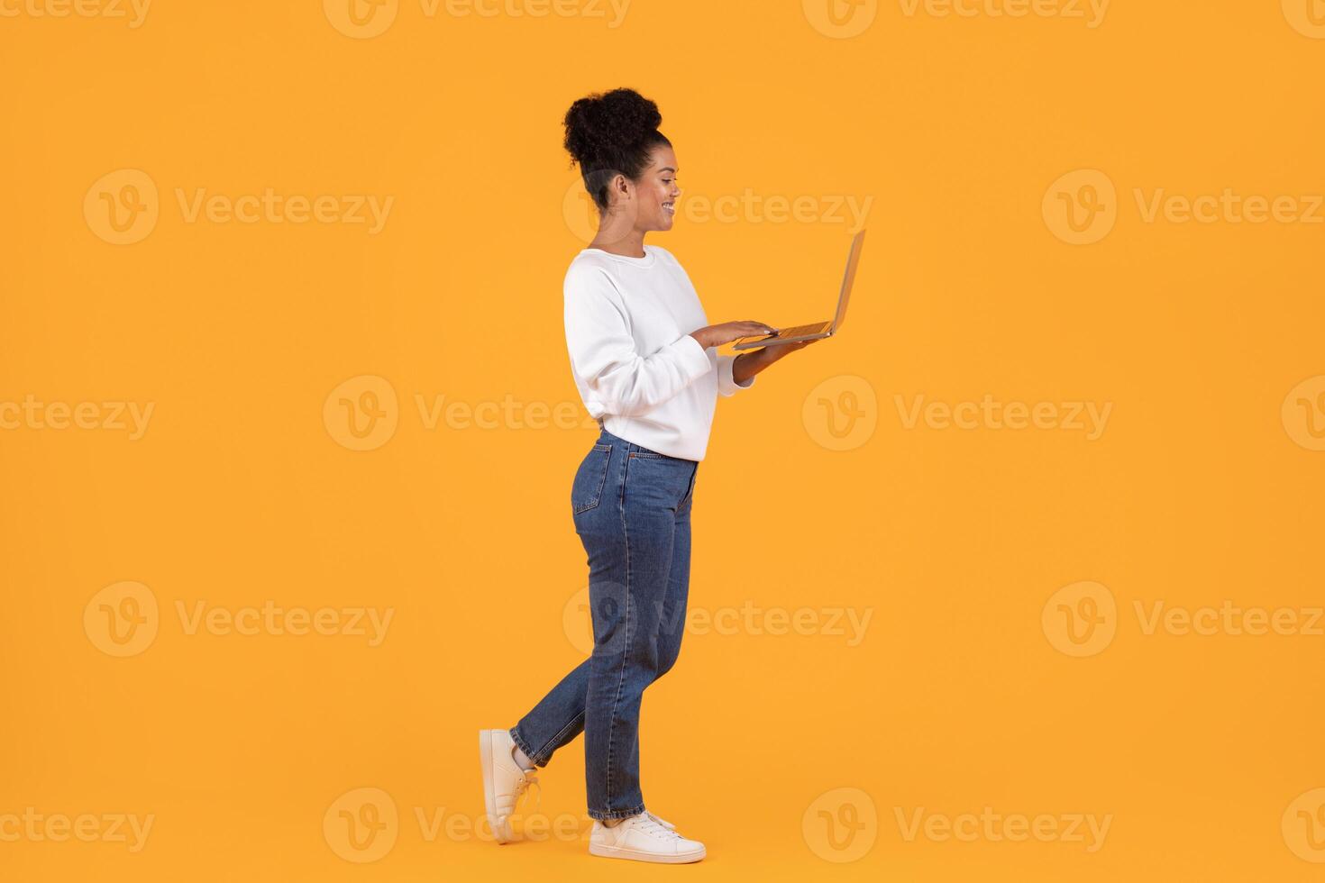 A young woman walks forward while looking at and using her laptop. She is wearing a white sweater, blue jeans, and white sneakers. She is smiling as she walks on a bright yellow background. photo