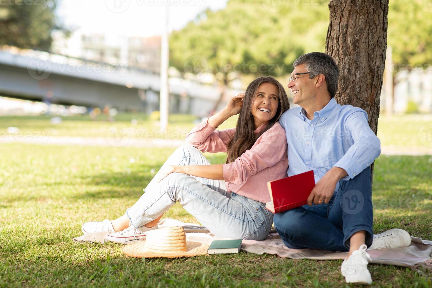 A couple sits under a tree in a park, sharing smiles and laughter while reading a book. The day is bright, showcasing a relaxed moment in nature together. photo