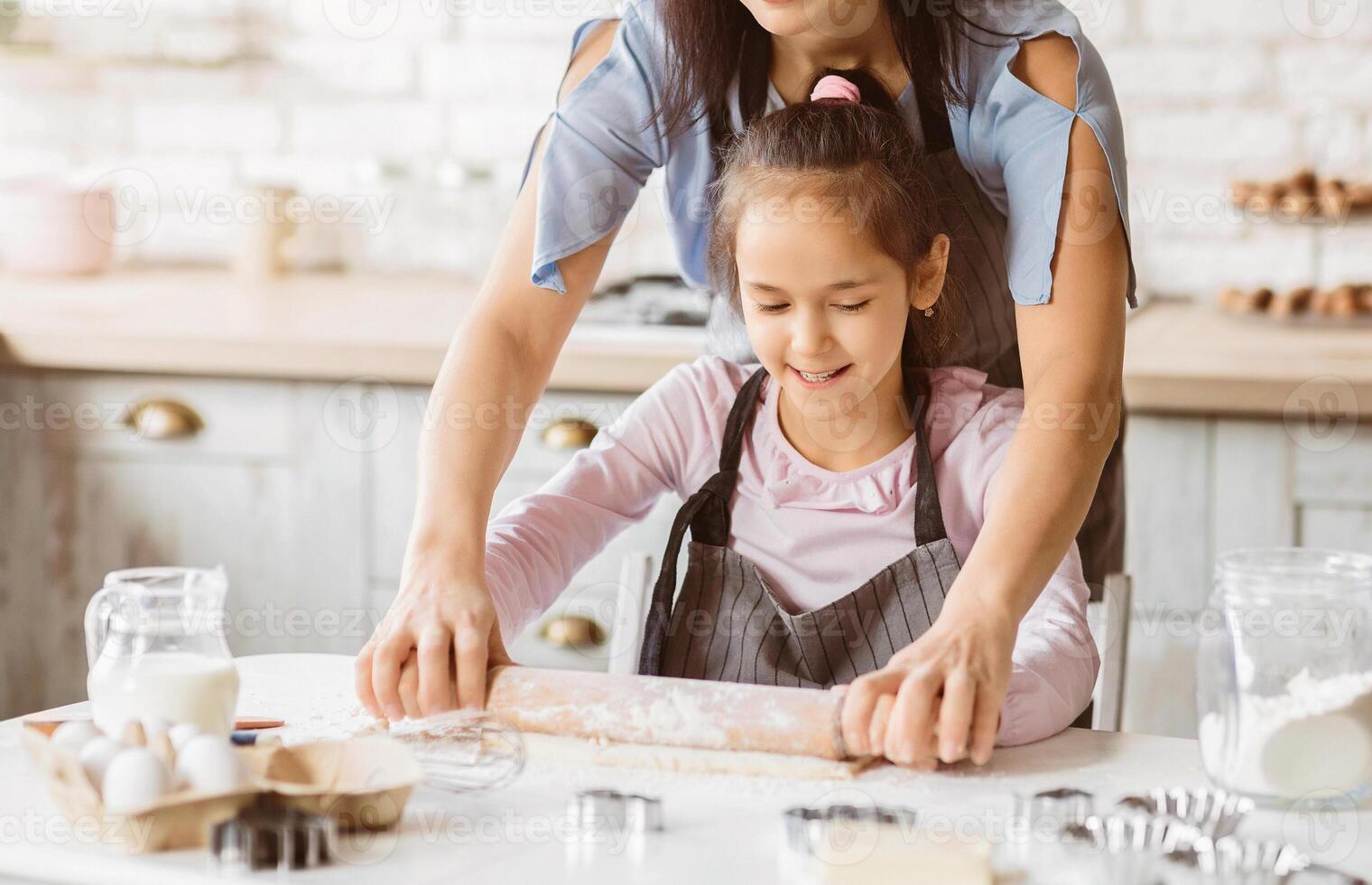 A young girl is learning to bake with her mother in the kitchen. She is using a rolling pin to flatten dough while her mother guides her hand. There are baking ingredients and tools on the table photo