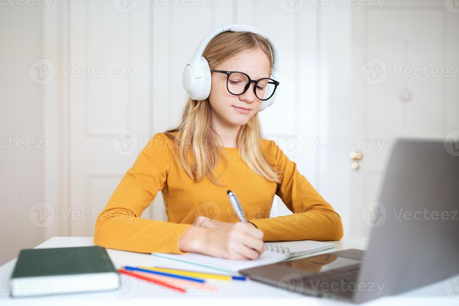 A teen girl, wearing glasses and headphones, sits at a desk with a laptop in front of her. She is writing in a notebook while looking at the screen. photo