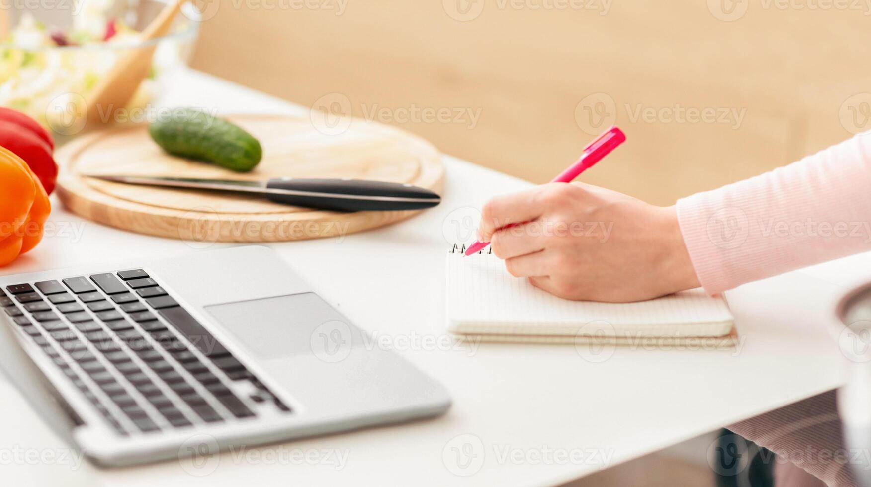 Cropped of lady is sitting at a desk, writing on a notebook next to a laptop. Woman is focused on her work, with pens and papers scattered around the workspace. photo