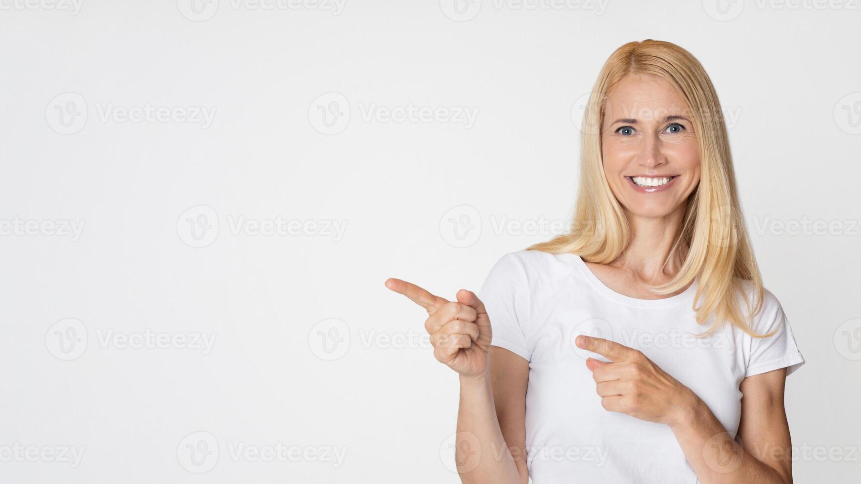 A woman with long blonde hair is smiling and pointing to the copy space in front of a plain white background. She is wearing a simple white t-shirt and has a friendly expression on her face. photo