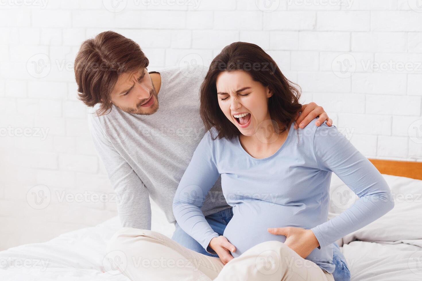 Prenatal contractions. Worried husband comforting pregnant wife that suffering from acute pain on bed at home, she is giving birth, copy space photo