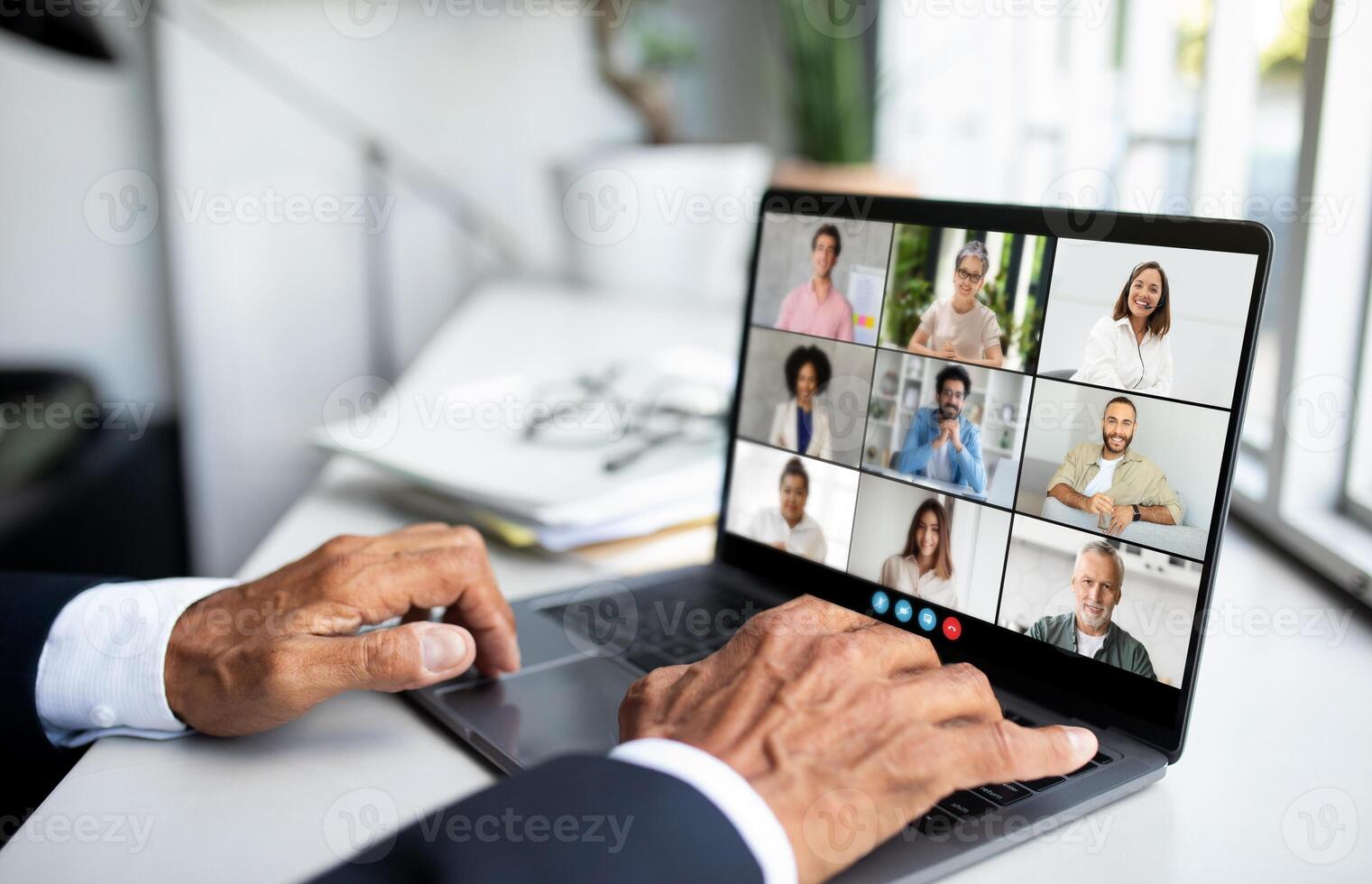 Unrecognizable man is sitting in front of a laptop, engaging in a call with a group of people displayed on the screen. He appears focused and attentive as he interacts with his virtual audience. photo