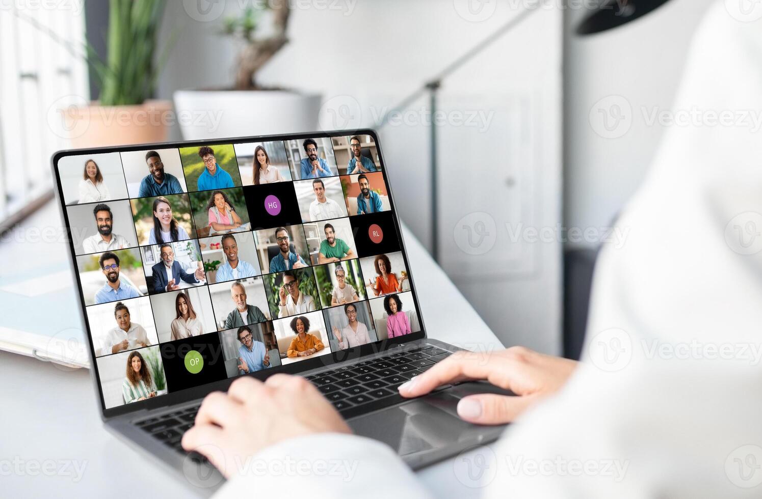 A woman is seated in front of a laptop, engaging with a group of people visible on the screen. She appears focused and attentive, possibly in a virtual meeting or online conference, cropped photo