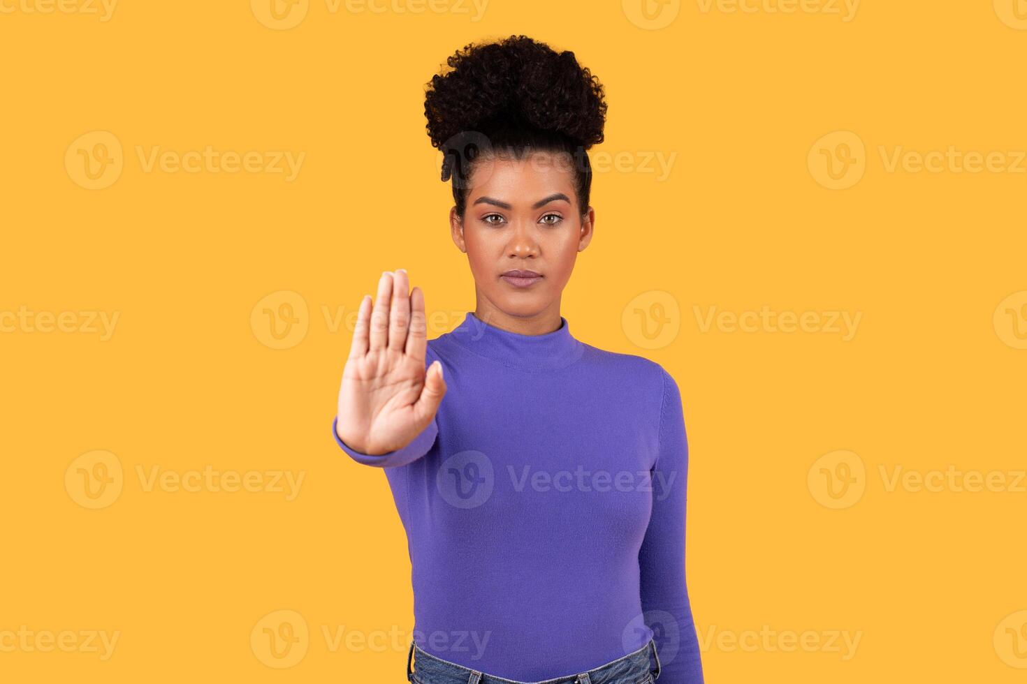 This image shows a woman with dark curly hair wearing a purple shirt and jeans. She is standing in front of a yellow background, holding her hand up in a stop gesture. photo