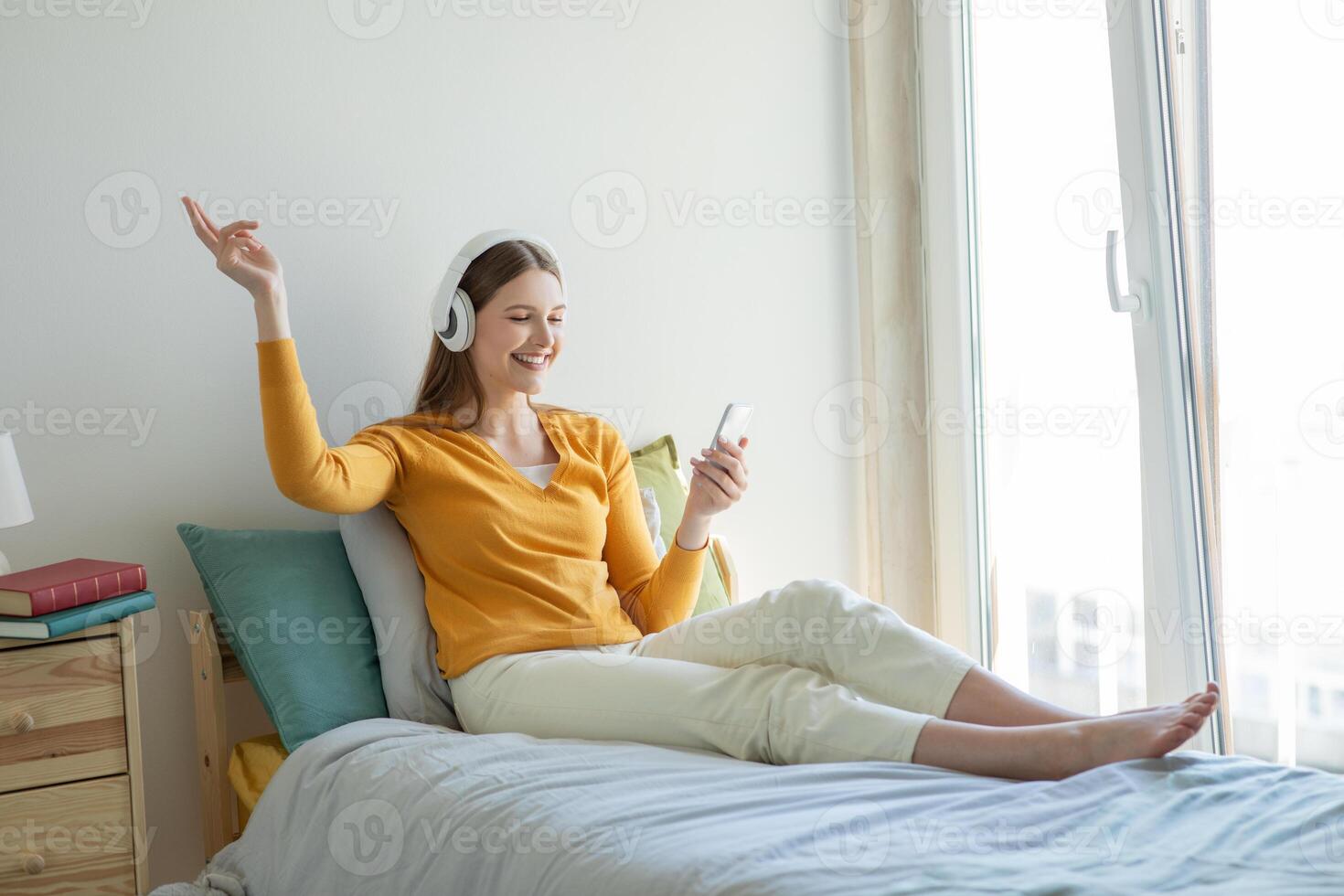 A girl is lounging on her bed, enjoying music through her headphones. She is smiling, relaxed, and appears to be in a light, airy room with natural sunlight streaming in through a large window. photo