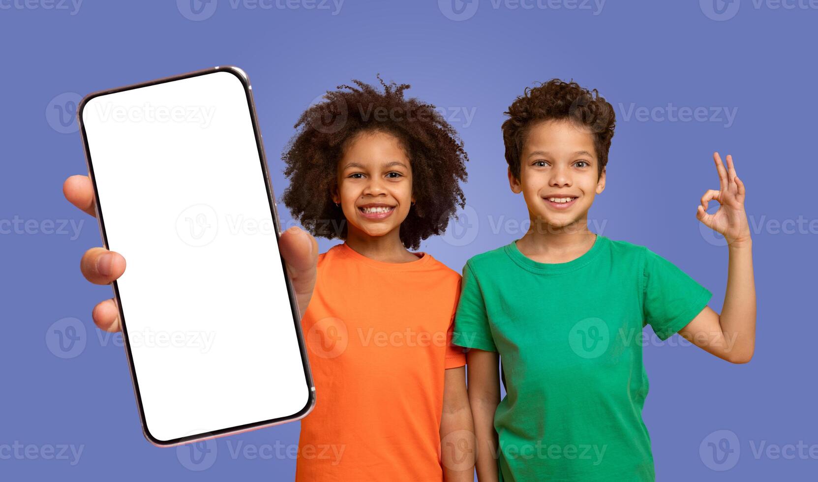Two black kids, a girl in an orange shirt and a boy in a green shirt, are smiling and holding a smartphone with a blank white screen. The boy is showing the okay hand gesture. photo