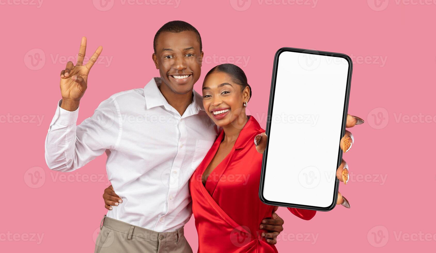 A happy, young black couple stands in front of a pink background. The man making a peace sign with his right hand. The woman holding a smartphone with a blank white screen photo