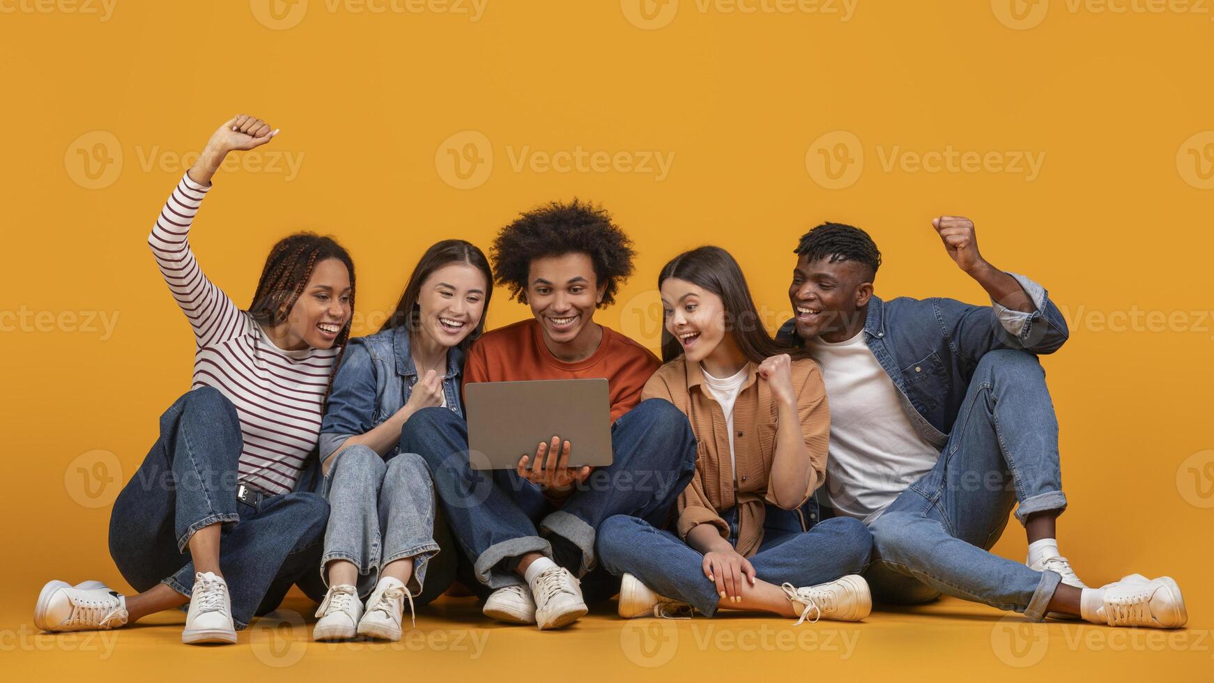 A group of five diverse friends sit on a yellow background, looking at a laptop and celebrating. They appear excited and happy, with smiles on their faces and arms raised in the air photo