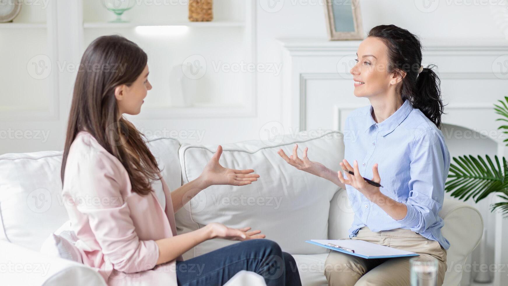 Two women are seated on a comfortable white sofa having an engaging conversation. One woman appears to be a therapist holding a clipboard, while the other, dressed casually, seems to be the client. photo