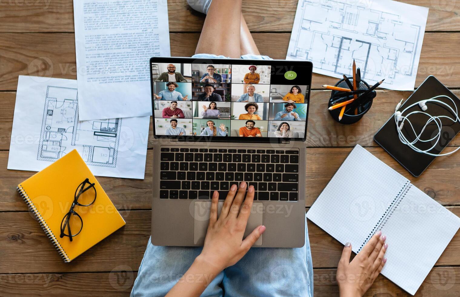 A woman is sitting on a wooden floor, using a laptop to attend a conference meeting. The laptop is open, displaying a screen filled with 16 participants in a grid format. photo