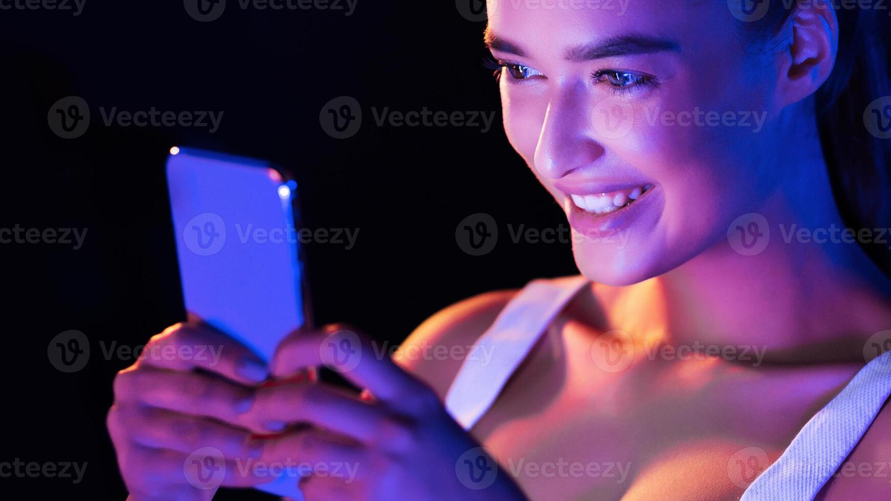 A young woman is seated, holding a smart phone in her hands and looking at the screen intently in a dark room at night. The glow from the phone illuminates her face as she interacts with the device. photo
